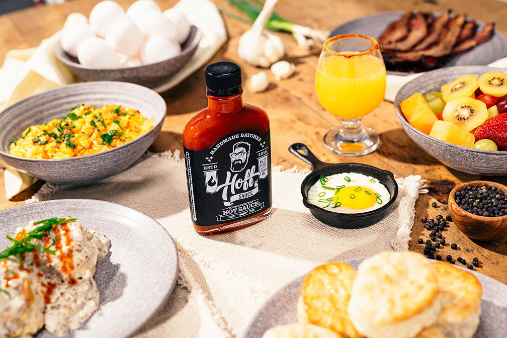 Handmade, small batch, aged Hoff & Pepper Louisiana fresh pepper hot sauce bottle on breakfast table with scrambled eggs, mimosas, biscuits and gravy, bacon, and a sunny-side-up egg in a skillet.